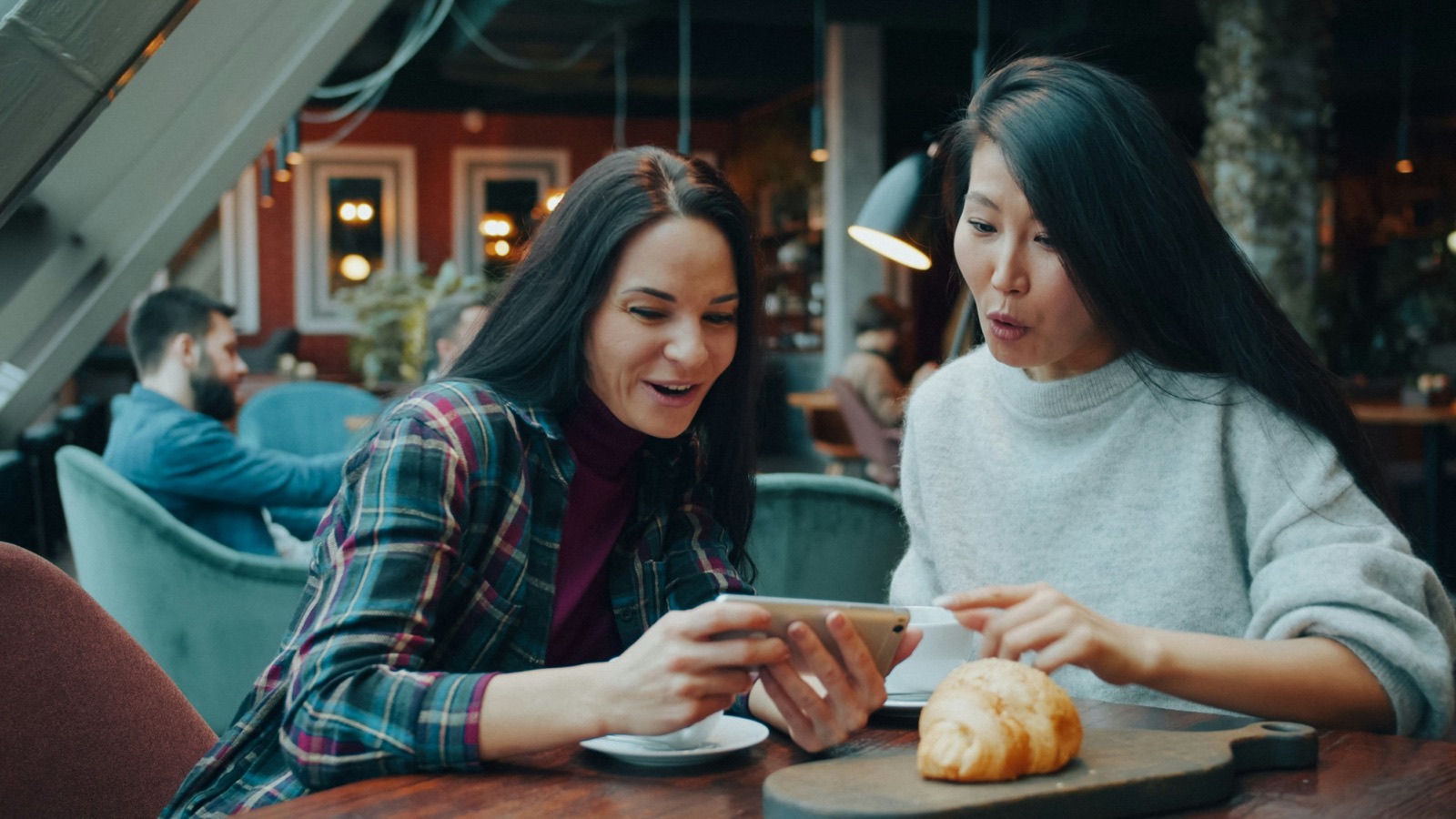 Two friends look at a phone together in a café, one of them surprised